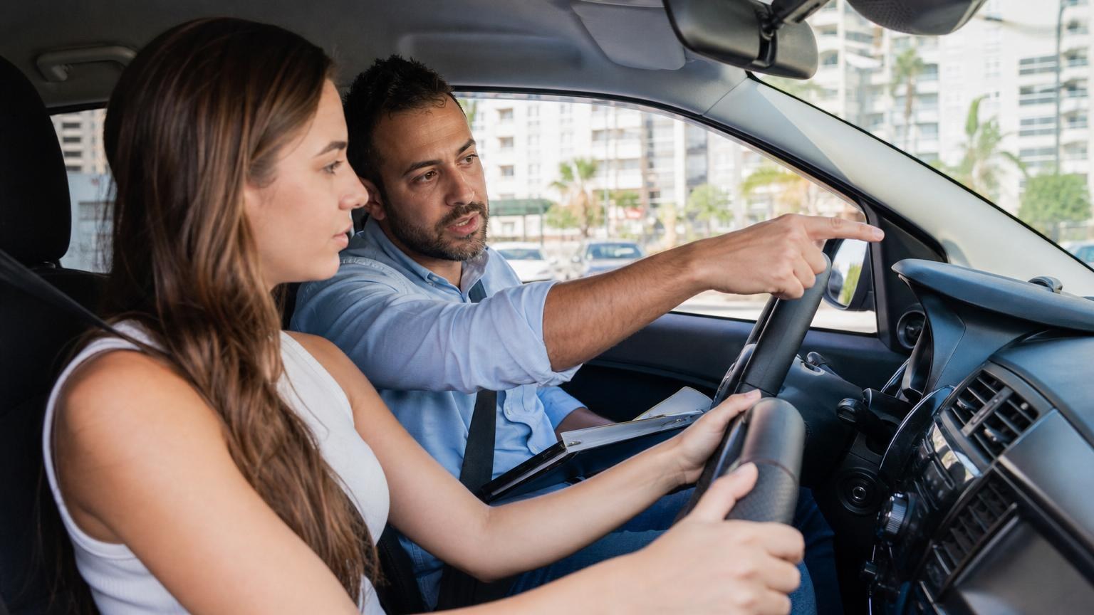 Male driving instructor pointing at the road while teaching a young woman behind the wheel of a car in a sunny city. Male driving instructor pointing at the road while teaching a young woman behind the wheel of a car in a sunny city.